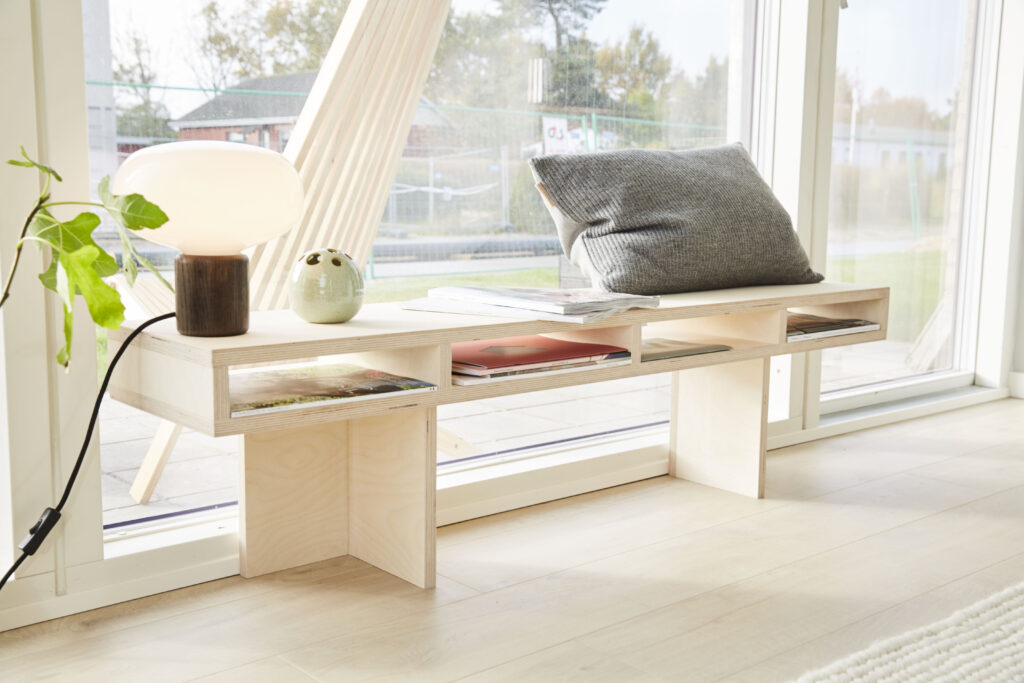 Fin nordisk bænk i krydsfinér med opbevaringsplads under. Pyntet med pude, lampe og blade.